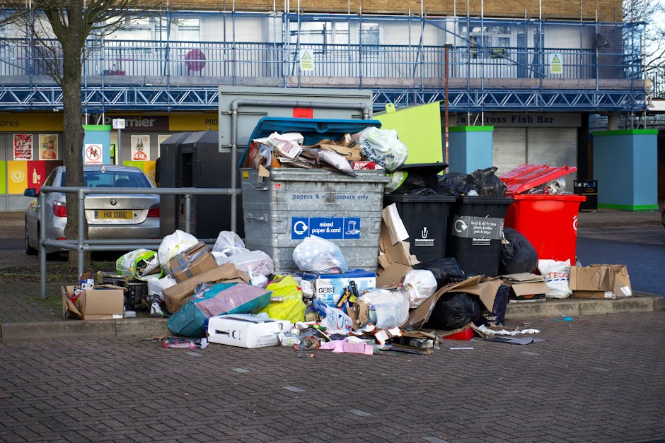 A large collection of mixed waste, including crumpled cardboard boxes, plastic bags, paper parcels, and various discarded packaging, is piled up outdoors on a paved area beside a small metal railing. The waste is densely packed around a central grey recycling bin labeled 'mixed paper & card,' with some rubbish spilling out onto the ground. Adjacent to the grey bin are a black rubbish bag, a red bin, and another black bin also filled with waste. In the background, there are retail units with closed shutters and signage, as well as a blue metal scaffolding structure covering the upper part of the building façade. A portion of a parked silver hatchback car can be seen on the left side, partially obscured by a tree. The scene appears to be in a commercial or shopping area, emphasizing the importance of waste collection and management services such as those provided by Waste Clearance Knightsbridge, with the overall environment showing signs of an overflowing waste area awaiting clearance or disposal, reflecting the need for professional rubbish removal assistance.