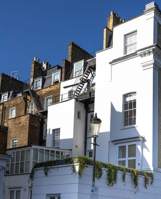 A close-up view of the upper floors of a row of terraced residential buildings in Knightsbridge, with the focus on a white exterior wall contrasting against adjacent brickwork. The white building features multiple windows with white frames, some with decorative trim, and a small balcony enclosed with glass panels on the lower level. Black metal fire escapes with ladders and railings are attached to the brick sections, extending vertically across several floors. A black street lamp with a traditional design stands on the pavement directly in front of the buildings, casting a shadow onto the white wall. The pavement is visible at the bottom of the image, with a small green vine growing over the edge of the balcony, adding a touch of greenery. The scene is brightly lit by natural sunlight against a clear blue sky, illustrating typical urban architecture and the type of properties where private or independent rubbish removal services might be employed to handle waste alongside building maintenance.