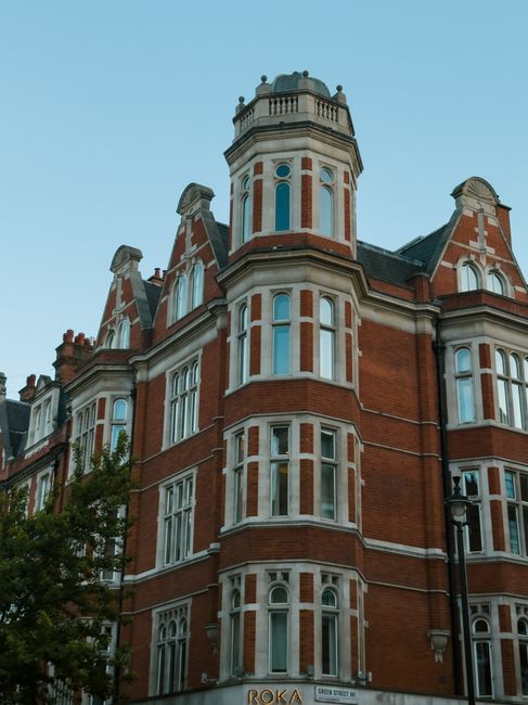 A tall, historic multi-storey red-brick building with decorative white window frames and ornate architectural details, including a rounded turret at the corner topped with a small, domed roof and balustrade. The structure features multiple bay windows spanning several floors, with some windows arched and others rectangular, all with white sashes and trim. The building's exterior shows a combination of smooth brickwork and decorative stone accents, with a prominent vertical emphasis created by the turrets and window alignment. In the foreground, a mature tree partially obscures the lower part of the building, and a black street lamp stands adjacent to it. The scene is illuminated by natural daylight, with a clear blue sky overhead, emphasizing the building's historical character and craftsmanship. The setting appears residential or commercial within an urban area, consistent with private property in a city environment, likening the scene to alternative waste handling options such as private rubbish collection at similar properties.