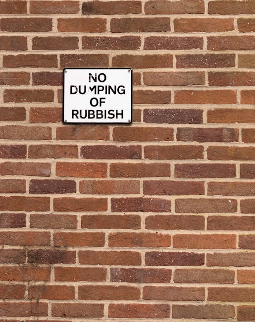 A rectangular metal sign with a white background and black text attached to a red brick wall. The sign reads 'NO DUMPING OF RUBBISH' with the word 'DUMPING' partially obscured due to a portion of the sign being bent or damaged. The brick wall features reddish-brown bricks with darker and lighter shades, arranged in a horizontal pattern with visible mortar joints, and extends across the entire background of the image. The lighting appears natural, creating slight shadows on the bricks and sign, and the scene likely depicts an exterior wall where waste management or rubbish removal regulations are displayed, aligning with the context of private waste handling or alternative collection methods. Waste Clearance Knightsbridge might use such signage to discourage illegal dumping and promote responsible rubbish disposal in the local area.
