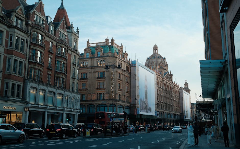 A busy street scene in Knightsbridge featuring a row of historic mixed-use buildings with ornate brick and stone facades, large windows, and decorative architectural details. The central building has a notable green-domed roof and elaborate cornices, with a large billboard display mounted on its front. To the right, a modern glass-fronted building with a blue horizontal awning extends partially into the frame. The street is filled with parked cars, including a classic red double-decker bus positioned near the center, indicating an urban environment with active pedestrian and vehicular traffic. People dressed in various styles walk along the pavement, some entering shops or browsing storefronts. The lighting suggests early evening, with a slightly overcast sky providing diffuse, natural light that highlights the textures and colours of the buildings and vehicles, evoking a typical scene of central London with its characteristic blend of historic architecture and modern retail spaces, aligning with the context of local waste management and rubbish clearance services in a prominent commercial area.