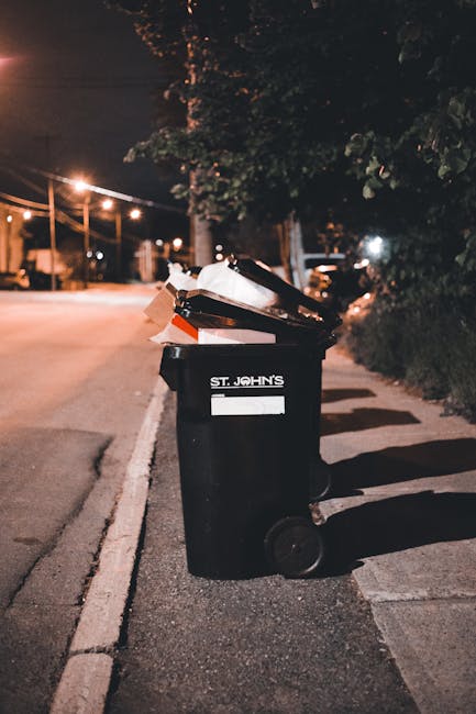 A black wheeled rubbish bin labeled 'St. John's' is positioned on a sidewalk next to a curb in an evening urban setting. The bin's lid is open, revealing a mixture of mixed waste including cardboard, paper, and plastic refuse. The surface of the bin appears textured and slightly worn, with some items displacing outward from the opening, indicating recent disposal. In the background, streetlights cast a warm glow, illuminating adjacent trees with dark, leafy foliage and a quiet street scene with parked vehicles and architecture partially visible in the distance. The environment suggests a typical residential or mixed-use area where private waste collection services, such as those provided by Waste Clearance Knightsbridge, might operate as an alternative to municipal rubbish collection. Shadows are cast by the bin and nearby objects, creating a sense of depth, while the night-time atmosphere underscores the importance of proper rubbish disposal in urban waste management practices.