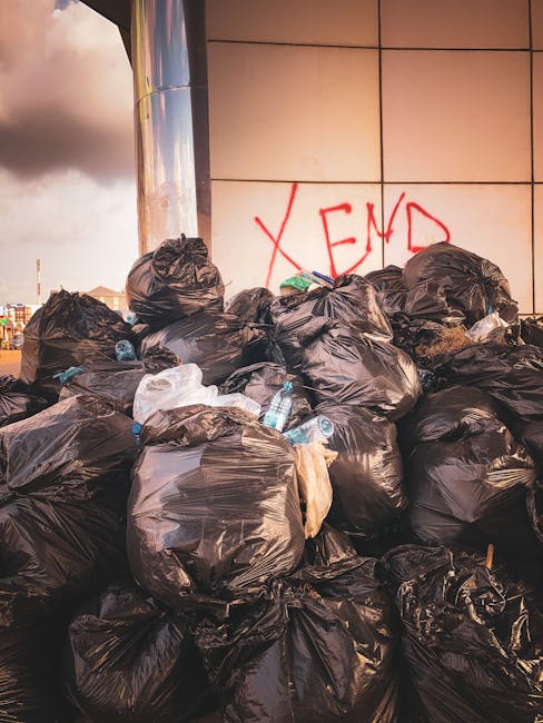A large pile of black rubbish bags, some slightly torn or bulging, is stacked against a modern building wall with a reflective surface in an urban setting. Several plastic bottles, mostly clear and partially crushed, are visible amongst the rubbish, with some protruding from the open or torn bags. The bags are stacked haphazardly, occupying most of the foreground, with the bags leaning on each other and some resting directly against the wall. Behind the pile, the wall features large, cream-colored tiles and red graffiti spelling 'XEND'. The environment appears to be an outdoor public area, possibly a pavement or alleyway adjacent to the building, with an overcast sky casting diffuse lighting over the scene. The presence of discarded bottles and the piled rubbish suggests an instance of private or independent waste disposal, aligning with services like those provided by Waste Clearance Knightsbridge for efficient rubbish removal and on-site clearance solutions.
