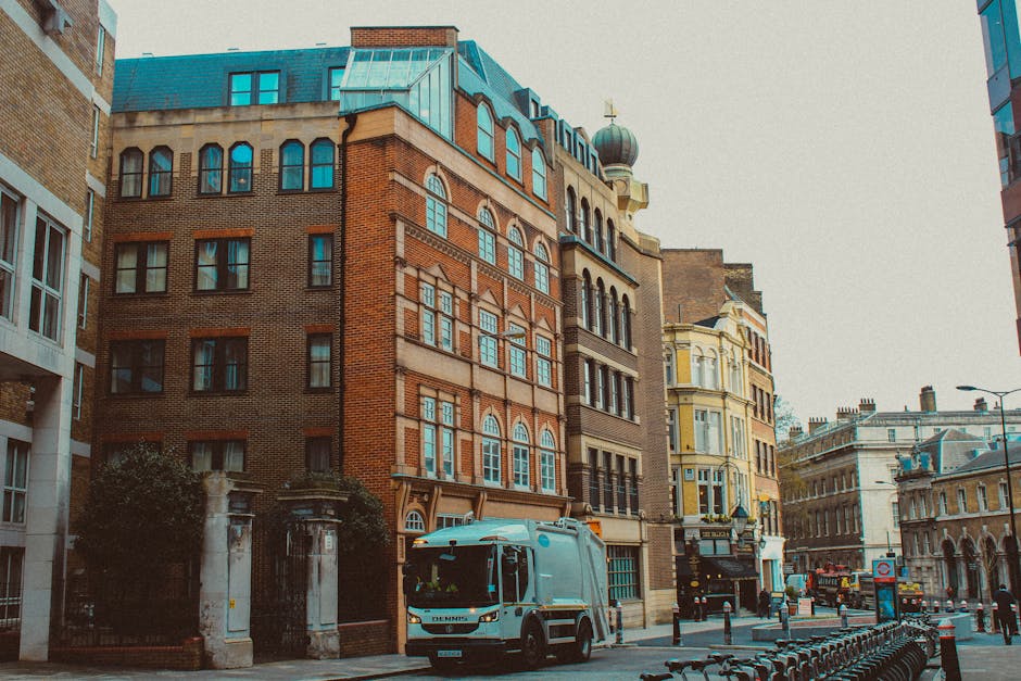 On a city street in a historic area, a grey waste collection truck is parked near a sidewalk adjacent to a row of multi-story buildings with diverse architectural styles. The foreground features the front of the truck with visible side mirrors and a front grille, positioned to load nearby rubbish or waste materials. To the left, there is an iron gate with stone pillars, suggesting an entrance to a private property or courtyard. The buildings display various facade finishes: one with exposed red brick and rectangular sash windows, another in a light yellow color with ornate window frames and a rounded turret with a dome roof topped by a decorative finial. The street environment includes bollards, parked bicycles, and modern street lighting, with a clear sky providing diffuse natural light that highlights the textures of the brickwork and architectural details. Waste Clearance Knightsbridge offers private waste management, demonstrated here by the presence of the collection vehicle ready for rubbish removal or on-site clearance, suitable for independent disposal needs outside standard council services.