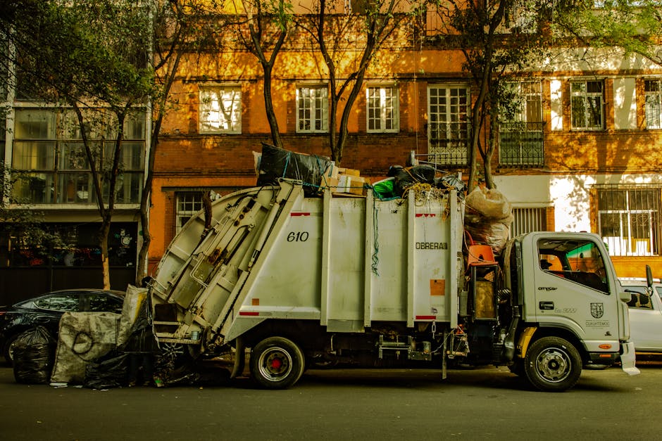A waste collection truck operated by Waste Clearance Knightsbridge is parked on a city street in front of a brick residential building with multiple windows and balconies. The truck is white with grey and black accents, featuring a large enclosed compactor at the rear that is tilted slightly upward, indicating it is either loading or ready to unload rubbish. The compactor is filled with various waste materials, including cardboard boxes, plastic bags, and miscellaneous debris, some of which are spilling over the edges. The side of the truck displays the company name and service category related to rubbish removal. In front of the truck, several black garbage bags are gathered on the tarmac pavement, some leaning against a small vehicle. The environment is shaded by trees whose branches and leaves cast dappled sunlight on the building façade and street, contributing to a calm, urban waste management scene focused on private rubbish collection and on-site clearance services.