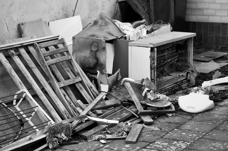 A pile of discarded household debris and rubbish is accumulated on a paved outdoor area next to a brick wall. The collection includes wooden pallets, which are leaning against the wall and partially broken, along with fragments of wood, torn fabric, and plastic debris scattered across the ground. Among the debris, there is an old piece of furniture with a damaged and exposed interior, possibly a broken cabinet or small appliance with its door and panels missing. A generic white plastic container and several pieces of torn foam or insulation are also visible. The environment suggests a site for waste clearance, with visible dirt and signs of neglect. This scene highlights a typical example of household waste that might be managed through private rubbish removal services such as those offered by Waste Clearance Knightsbridge, providing an example of on-site clearance needs for bulky or heavy waste materials in an urban setting. Lighting appears to be natural, casting some shadows over the clutter, emphasizing the disorderly nature of the rubbish pile.