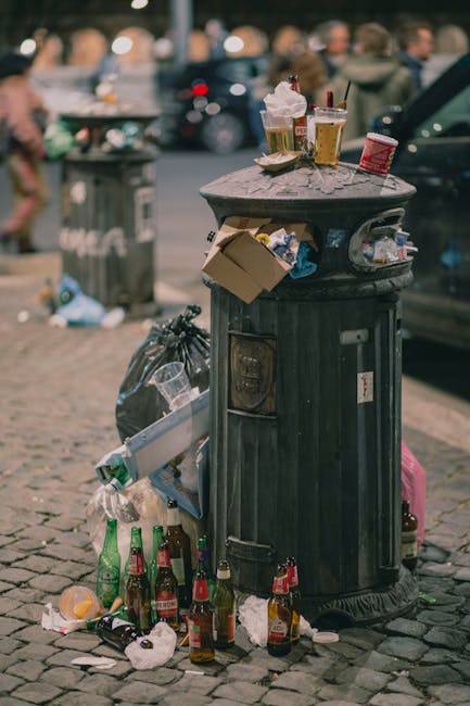 A black cylindrical public rubbish bin with a domed lid is situated on a cobbled street, serving as an overflowing litter receptacle in an urban environment. Sitting on top of the bin are remnants of food and beverage containers including a crumpled white paper napkin, disposable coffee cups with plastic lids, and a small soda can, all displaying signs of recent use. Attached to the side of the bin is a small food container or wrapper. Around the base of the bin, there is a collection of scattered waste, notably several glass bottles in red, green, and clear varieties, some with labels still attached, alongside crumpled paper and plastic wrappers. A black plastic trash bag is tied nearby, leaning against the side of the bin, alongside a biohazard waste bag in pink. In the blurred background, other trash receptacles and a few pedestrians are visible, illustrating an area with recent littering that could benefit from waste management services. The scene emphasizes the importance of responsible rubbish disposal and the need for efficient private waste handling, aligning with the services provided by Waste Clearance Knightsbridge.